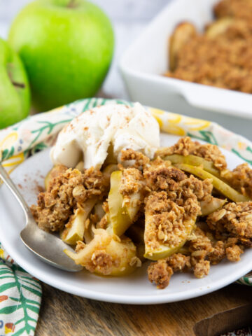 vegan apple crisp on a white plate with vegan ice cream with two Granny Smith apples in the background