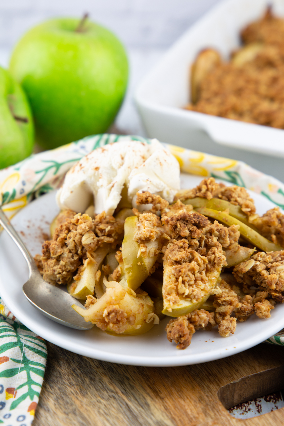 vegan apple crisp on a white plate with vegan ice cream with two Granny Smith apples in the background 