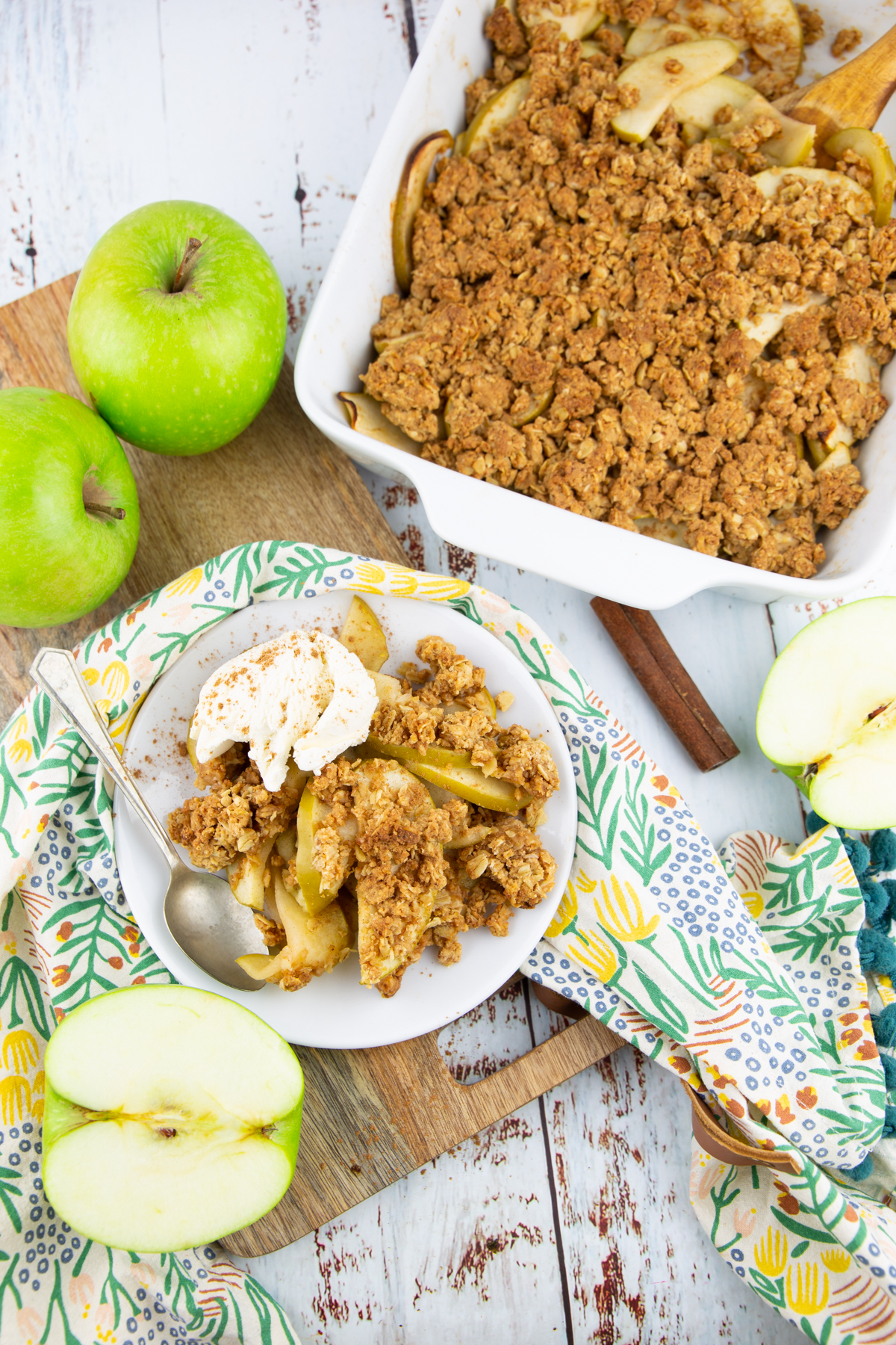 a white baking dish with apple crisp on a white wooden board with apples on the side 
