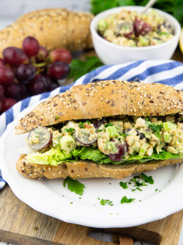 a white plate with a bun with vegan chicken salad on lettuce with grapes in the background