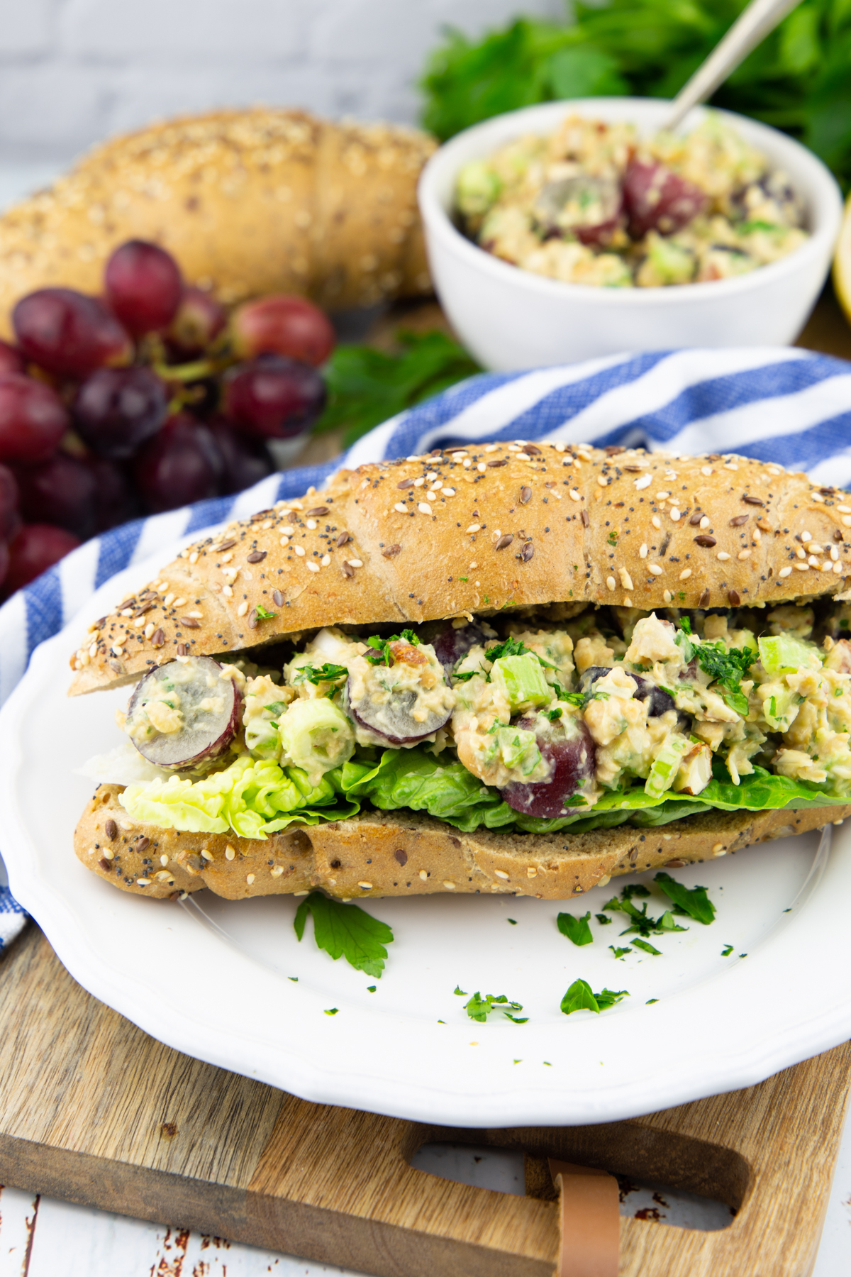 a white plate with a bun with vegan chicken salad on lettuce with grapes in the background 