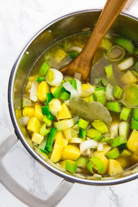 potatoes, leek, and vegetable broth in a pot with a wooden spoon on a marble countertop 