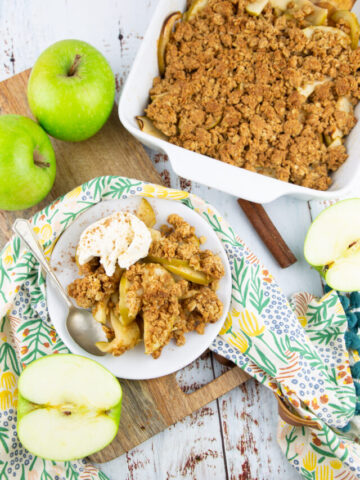 a white baking dish with apple crisp on a white wooden board with apples on the side