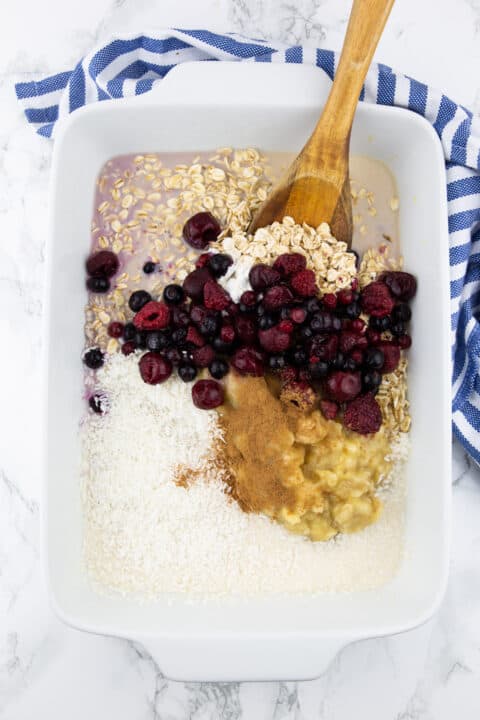 a white casserole dish with oats, berries, and plant-based milk with a wooden spoon 