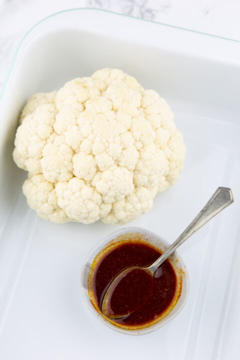 a head of cauliflower in a white baking pan with a small bowl of marinade on the side 