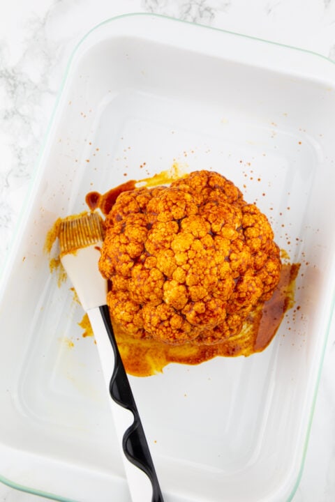 a head of cauliflower covered in marinade in a baking pan with a pastry brush on the side 