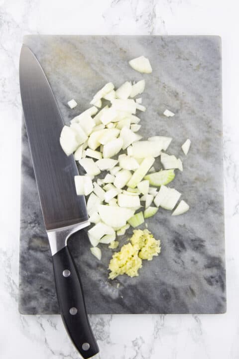 chopped onion and garlic on a marble chopping board with a large knife on the side 