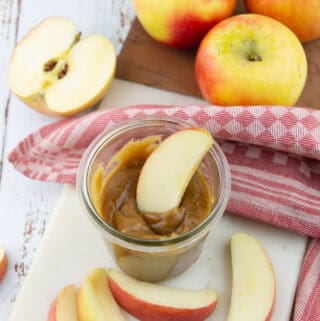 a glass jar with vegan caramel sauce with a sliced apple with more apples in the background