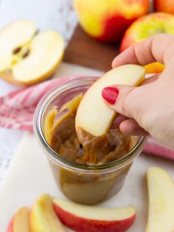 a hand dipping a slice of apple into a small glass with vegan caramel sauce
