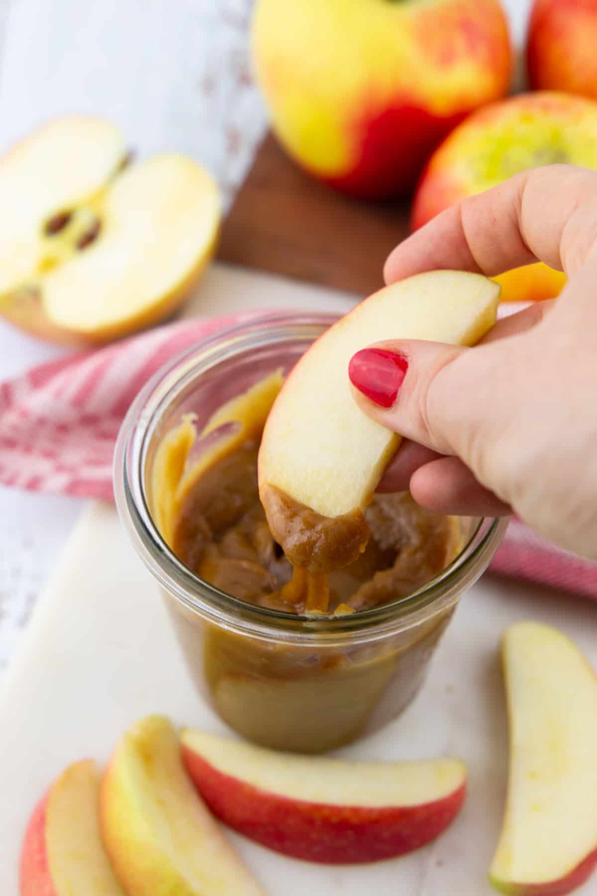 a hand dipping a slice of apple into a small glass with vegan caramel sauce 