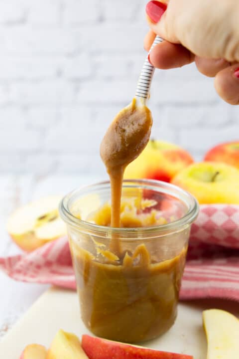 a hand holding a small spoon with vegan caramel sauce over a glass with more sauce with apples in the background