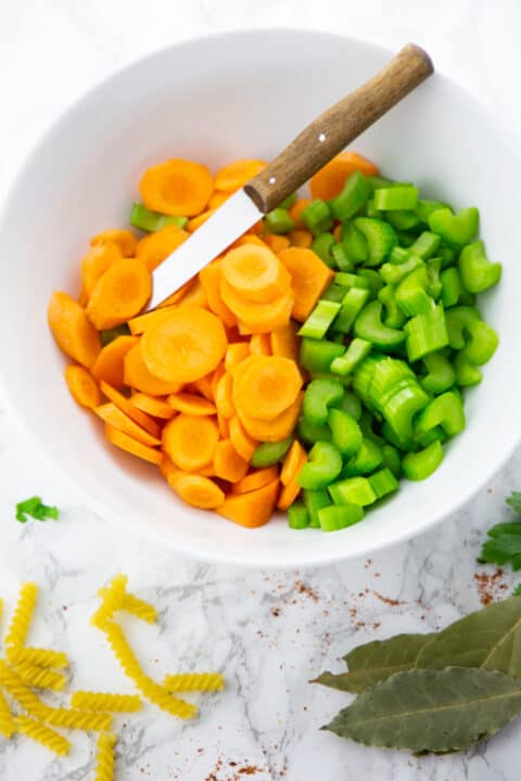 sliced carrots and celery in a white bowl with a knife on a marble countertop 