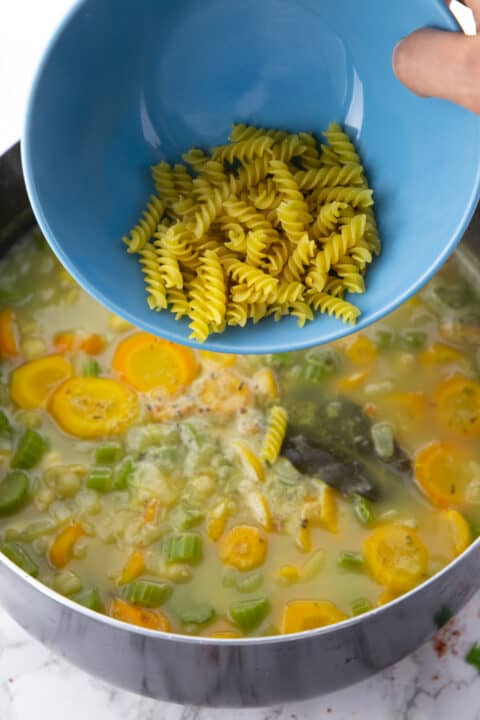 a hand pouring uncooked pasta into a large pot with vegetables and vegetable broth 
