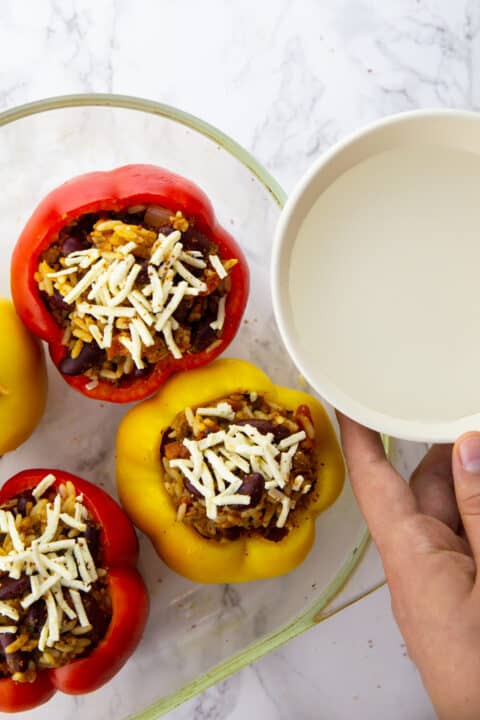 a hand pouring water into a casserole dish filled with stuffed peppers