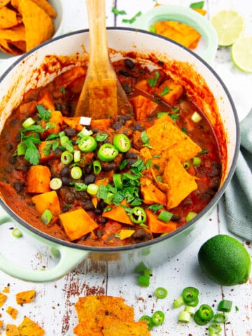 a green pot with sweet potato black bean chili on a white wooden board with limes and nachos in the background