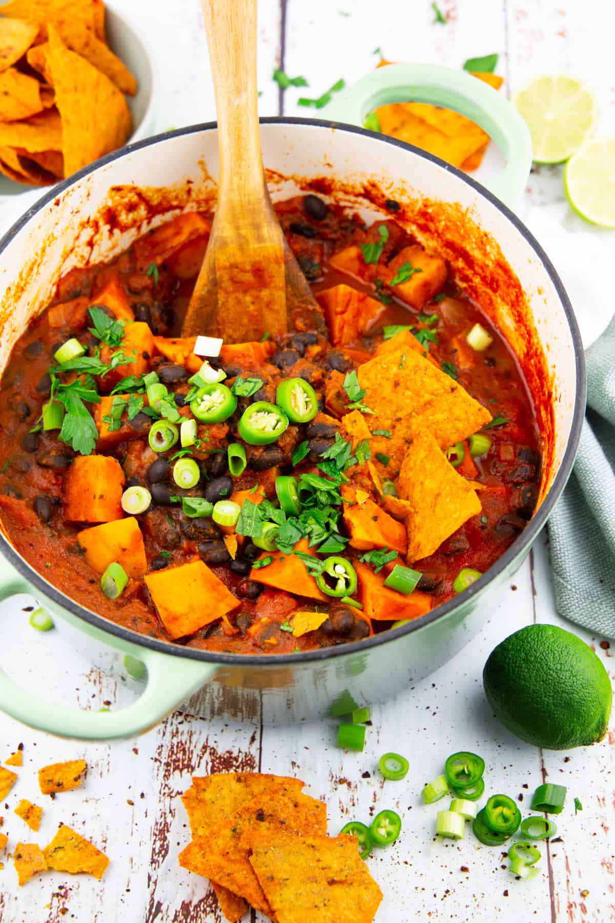 a green pot with sweet potato black bean chili on a white wooden board with limes and nachos in the background 