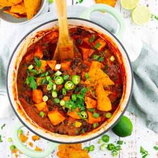 a green pot with sweet potato black bean chili on a white wooden board with limes and nachos in the background