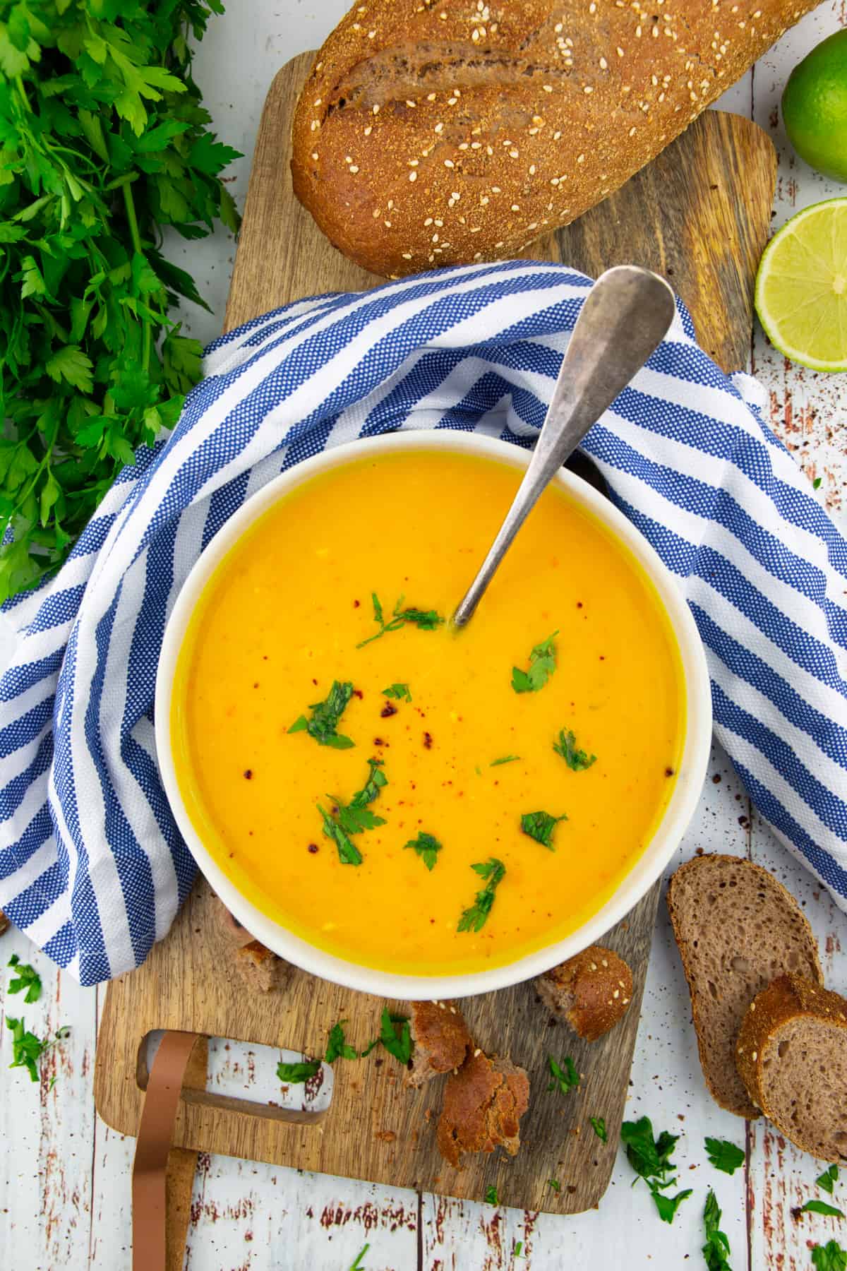 a bowl with sweet potato soup on a wooden board with a bunch of parsley and a loaf of bread in the background 