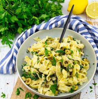 a grey bowl with tahini pasta on a wooden board with a bunch of parsley and lemons in the background