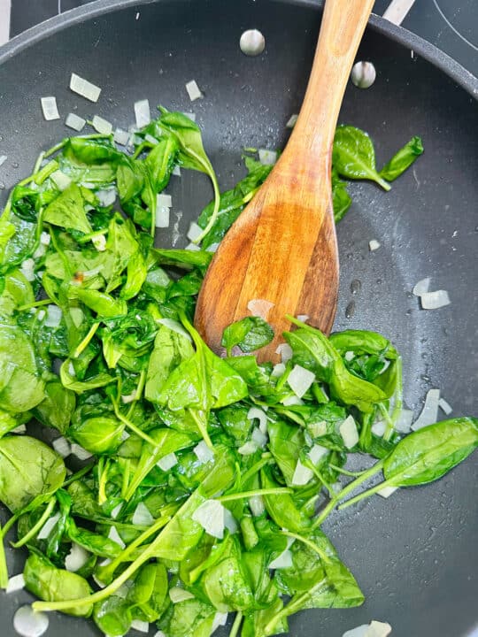 sautéd onion, garlic, and baby spinach in a black pan with a wooden spoon 