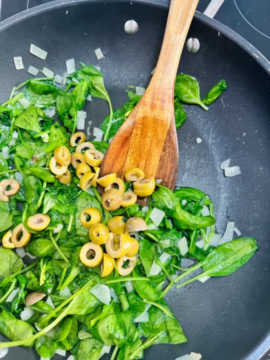 onion, garlic, baby spinach, and green olives in a black pan with a wooden spoon 