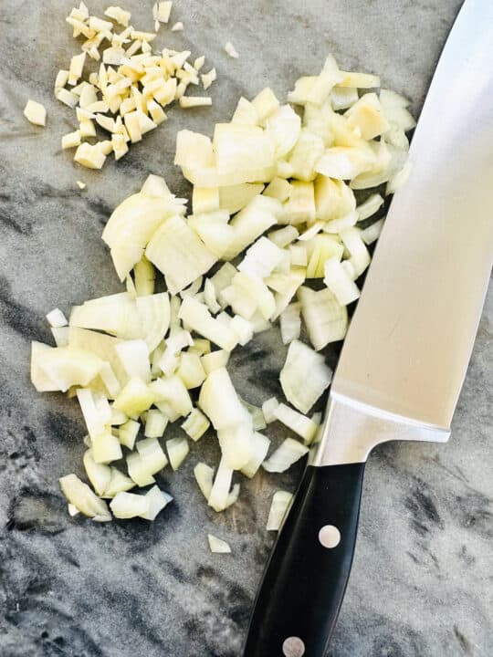 finely diced onion and garlic on a marble chopping board with a large knife 