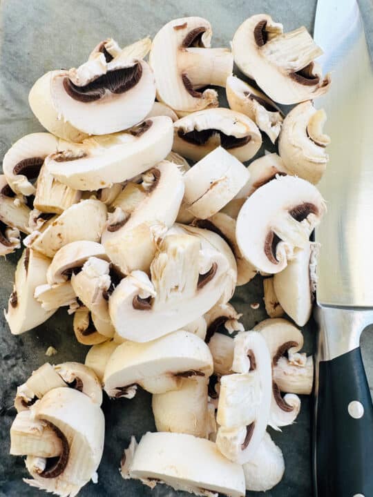 Sliced mushrooms on a marble chopping board with a large knife on the side 