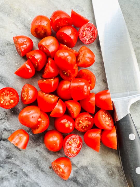 halved cherry tomatoes on a marble chopping board with a large knife on the side 
