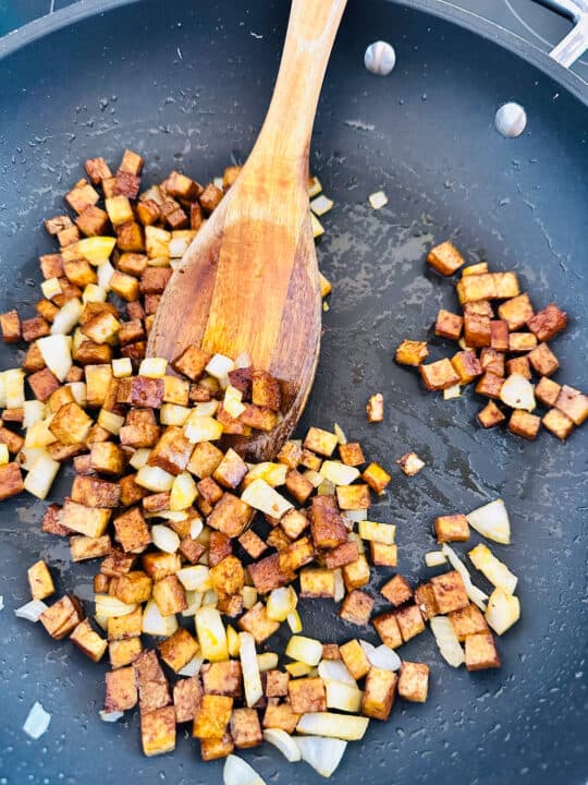 a black pan with pan-fried smoked tofu cubes and onion with a wooden spoon 