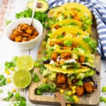 seven tempeh tacos on a wooden chopping board with a small white bowl with tempeh cubes and lime on the side