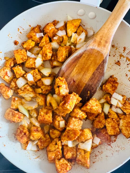 pan-fried tempeh cubes with sautéd onion in a white pan with a wooden spoon 