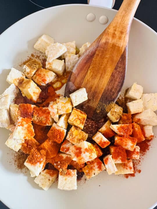 a white pan with tempeh cubes covered in spices with a wooden spoon 