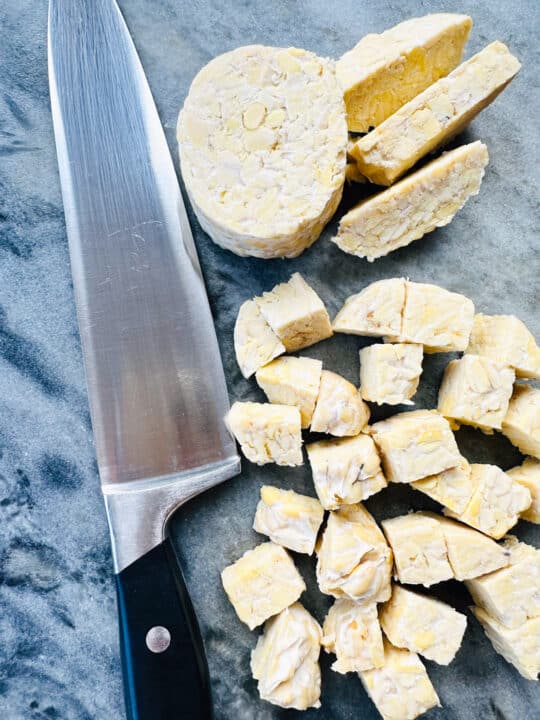 tempeh cut into cubes on a marble chopping board with a large knife on the side 