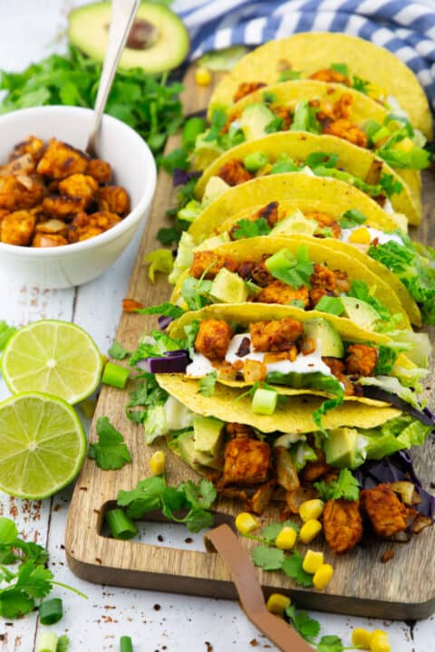 seven tacos filled with tempeh, lettuce, red cabbage, and avocado on a wooden chopping board with two lime halves on the side 