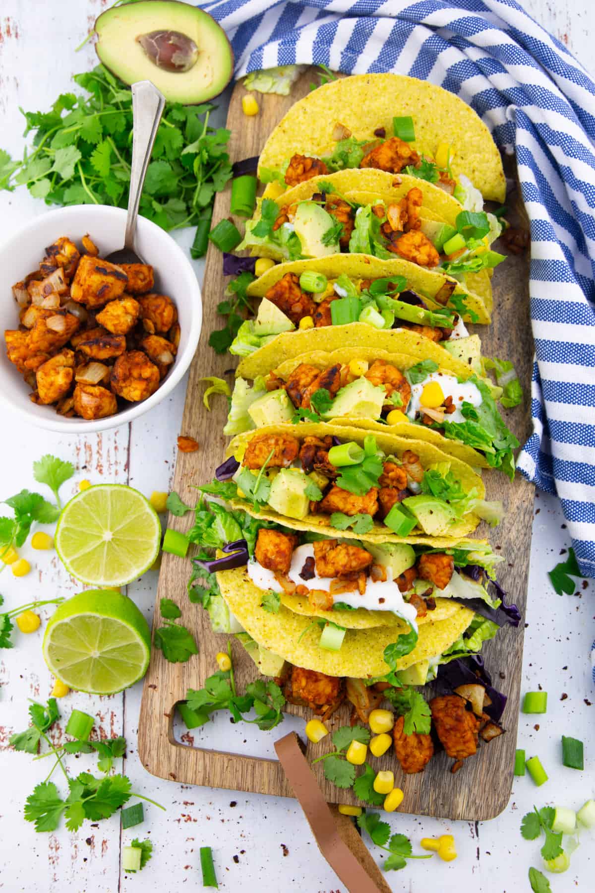 seven tempeh tacos on a wooden chopping board with a small white bowl with tempeh cubes and lime on the side 