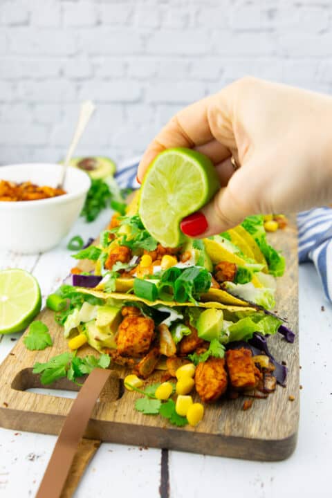 a hand squeezing a lime over tempeh tacos arranged on a wooden board 