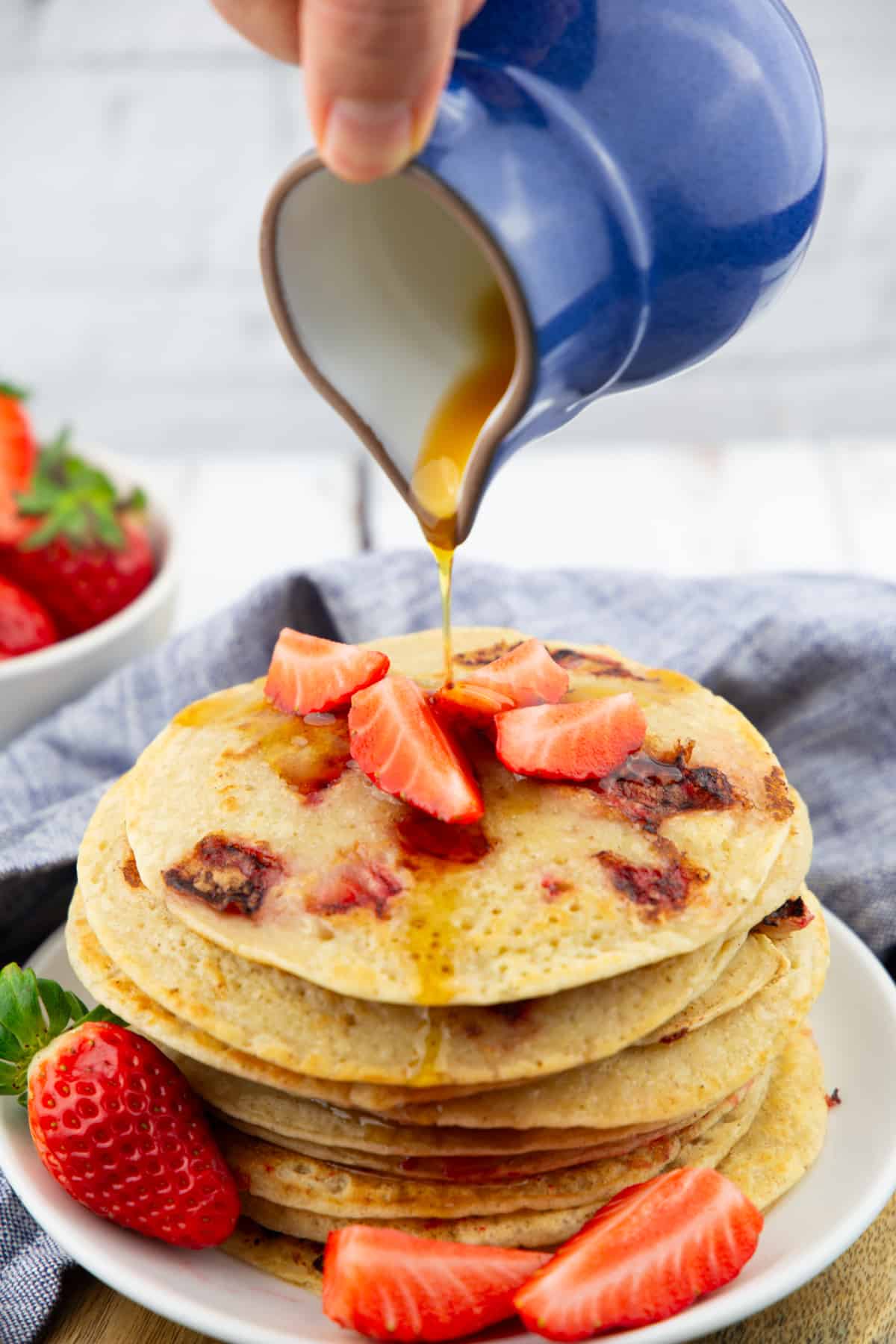 a stack of vegan pancakes with strawberries with a hand holding a blue small jug pouring maple syrup over the pancakes 