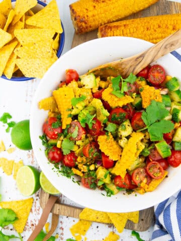 a white bowl with corn avocado tomato salad with a small bowl of nachos in the background and limes on the side