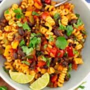 a grey plate with black bean pasta with bell pepper, lime wedges, and fresh cilantro on a wooden board