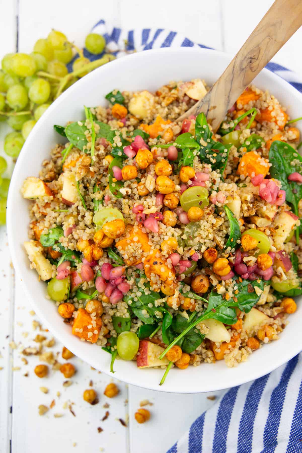 a white bowl with quinoa salad with sweet potato, spinach, and pomegranate on a white wooden board with grapes in the background