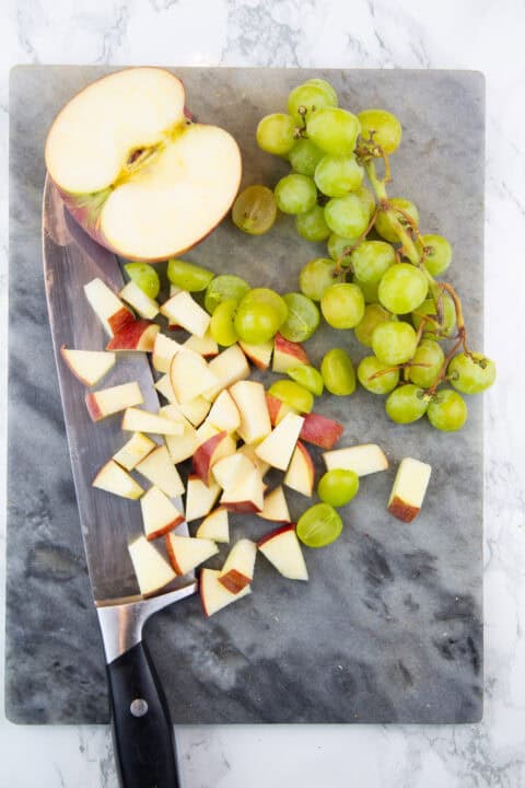 chopped apple pieces and green grapes on a marble chopping board with a large knife on the side