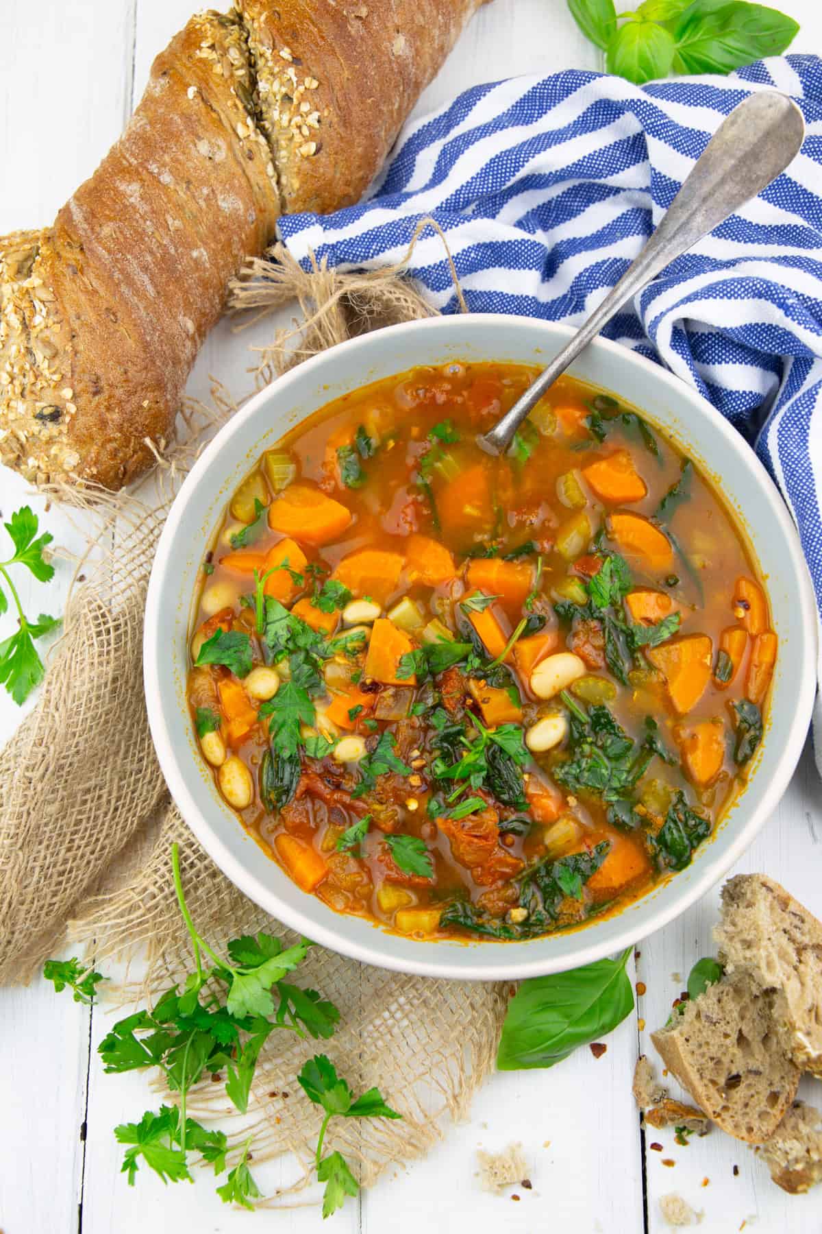 a grey bowl with Tuscan white bean soup with sun-dried tomatoes on a white wooden board with bread, parsley, and basil on the side