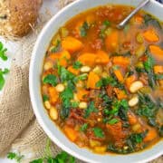 a bowl with Tuscan white bean soup with spinach on a white wooden board with bread in the background