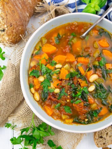 a bowl with Tuscan white bean soup with spinach on a white wooden board with bread in the background