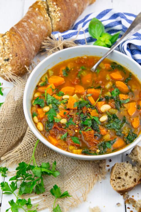 a grey bowl with Tuscan white bean soup on a white wooden board with a loaf of bread in the background
