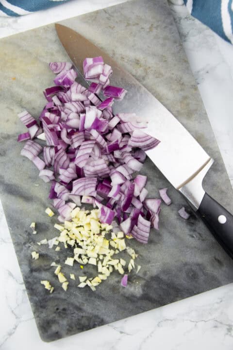 chopped red onion and garlic on a marble chopping board with a large knife on the side