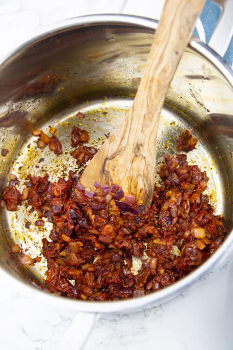 a pot with sautéed onion, garlic, sun-dried tomatoes, and tomato paste with a wooden spoon