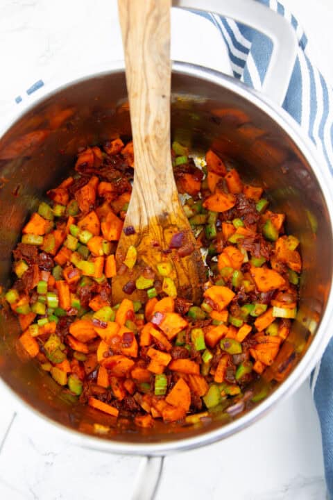 a pot with sautéed onion, chopped carrots, celery, and sun-dried tomatoes with a wooden spoon